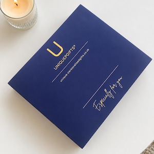 May include: A square navy blue gift box with gold lettering that reads UNIQUEPGIFTS, unique-personalisedgifts.co.uk, and Especially for you. A lit candle in a clear glass holder is positioned in the upper left corner of the frame.