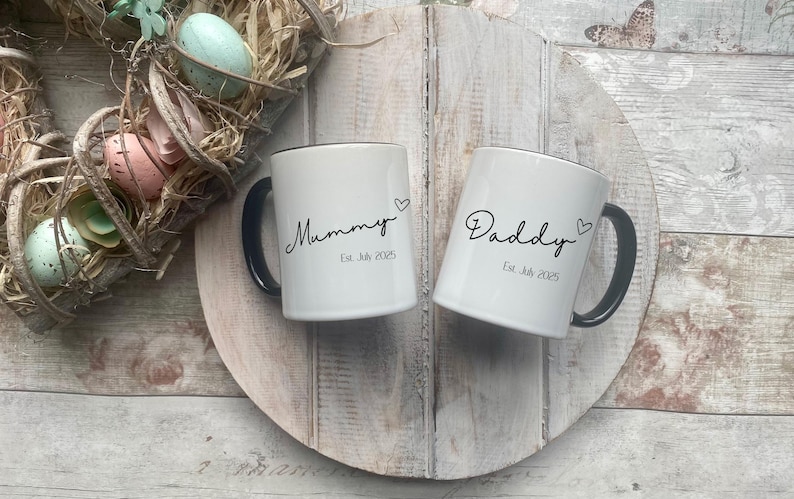 May include: Two white ceramic mugs with black handles and rims, inscribed with "Mummy" and "Daddy" in cursive, each with a heart symbol. The mugs are on a wooden surface, with an Easter wreath in the background.