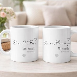 May include: Two white ceramic mugs with black text. One mug says "Soon To Be Mrs Smith ENGAGED 2016" and the other says "One Lucky Mr Smith ENGAGED 2016". Each mug features a heart symbol.