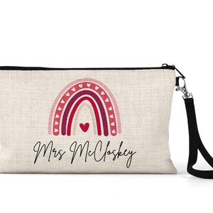 Pode incluir: Uma bolsa de maquilhagem de lona bege com um fecho de correr preto e uma correia preta. A bolsa apresenta um arco-&iacute;ris rosa, vermelho e roxo com cora&ccedil;&otilde;es brancos e o texto "Mrs. Mcloskey".