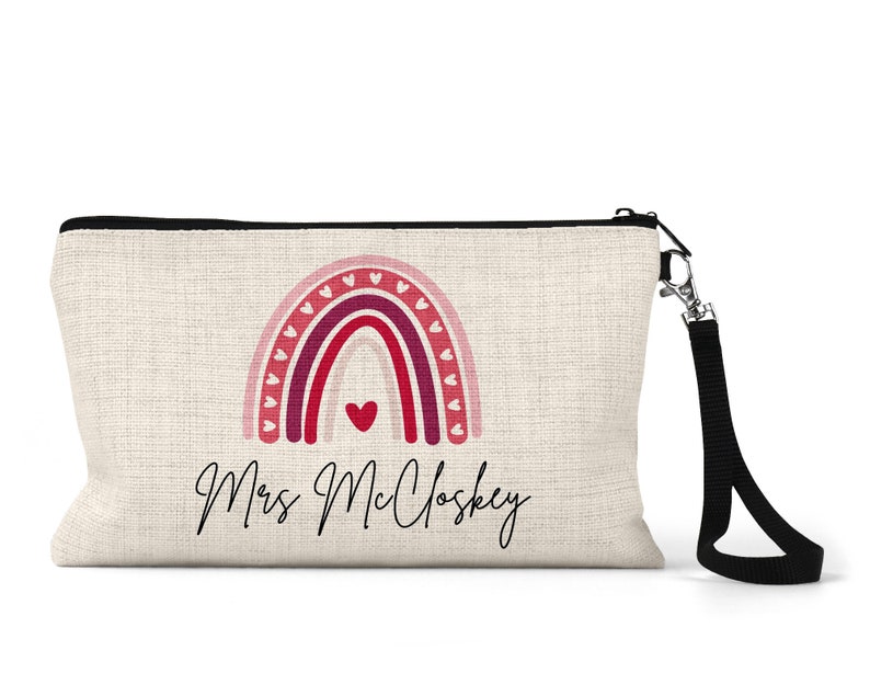 Pode incluir: Uma bolsa de maquilhagem de lona bege com um fecho de correr preto e uma correia preta. A bolsa apresenta um arco-&iacute;ris rosa, vermelho e roxo com cora&ccedil;&otilde;es brancos e o texto "Mrs. Mcloskey".