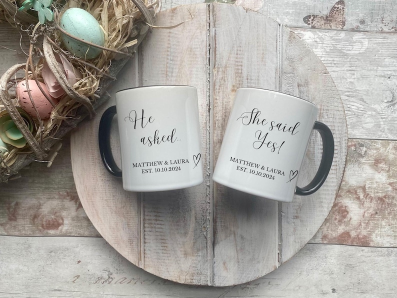 May include: Two white ceramic mugs with black handles and rims. One mug reads "He asked" and the other reads "She said Yes!" in elegant script. Both mugs include the names "MATTHEW & LAURA" and the date "EST. 10.10.2024".