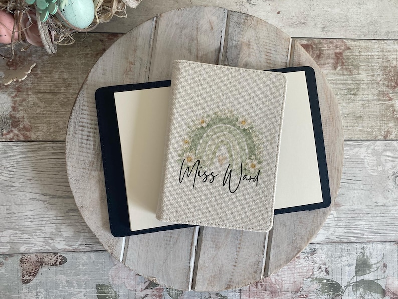 May include: A beige passport holder featuring a sage green rainbow graphic surrounded by white flowers and the text Miss Ward in black script. The holder is positioned on top of a black notebook with cream-coloured pages, displaying a textured fabric finish.
