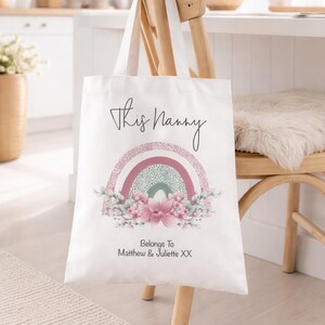 Op de afbeelding: Witte draagtas met de tekst "This Nanny" boven een regenboogontwerp in roze, groen en wit. De onderkant van de tas heeft de tekst "Belongs To Matthew & Juliette XX". De tas hangt aan een houten stoel.