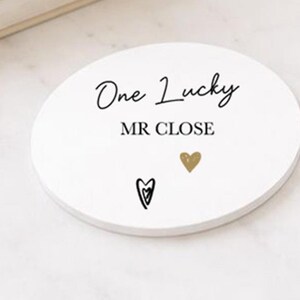 May include: A white, circular coaster featuring the text "One Lucky MR CLOSE" in black script. It also has a gold heart and a black heart design. The coaster is on a white surface.