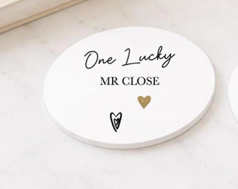 May include: A white, circular coaster featuring the text "One Lucky MR CLOSE" in black script. It also has a gold heart and a black heart design. The coaster is on a white surface.