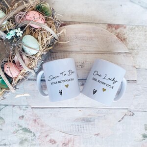 May include: Two white ceramic mugs with black text reading "Soon To Be Mrs Robinson" and "One Lucky Mr Robinson". Each mug features a gold heart and a black heart. The mugs are on a wooden surface.
