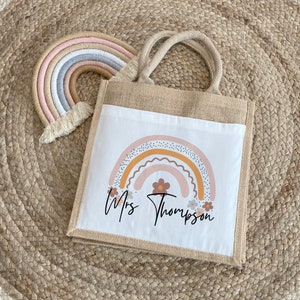May include: A white tote bag with a brown burlap handle. The bag has a printed design of a rainbow with flowers and the text "Mrs. Thompson".