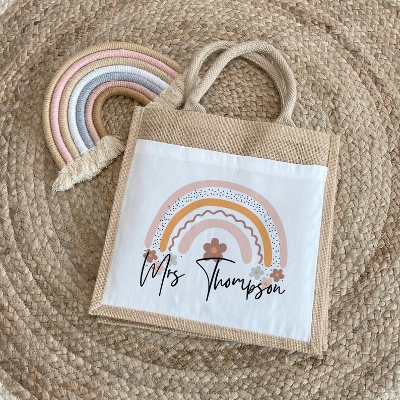 May include: A white tote bag with a brown burlap handle. The bag has a printed design of a rainbow with flowers and the text "Mrs. Thompson".