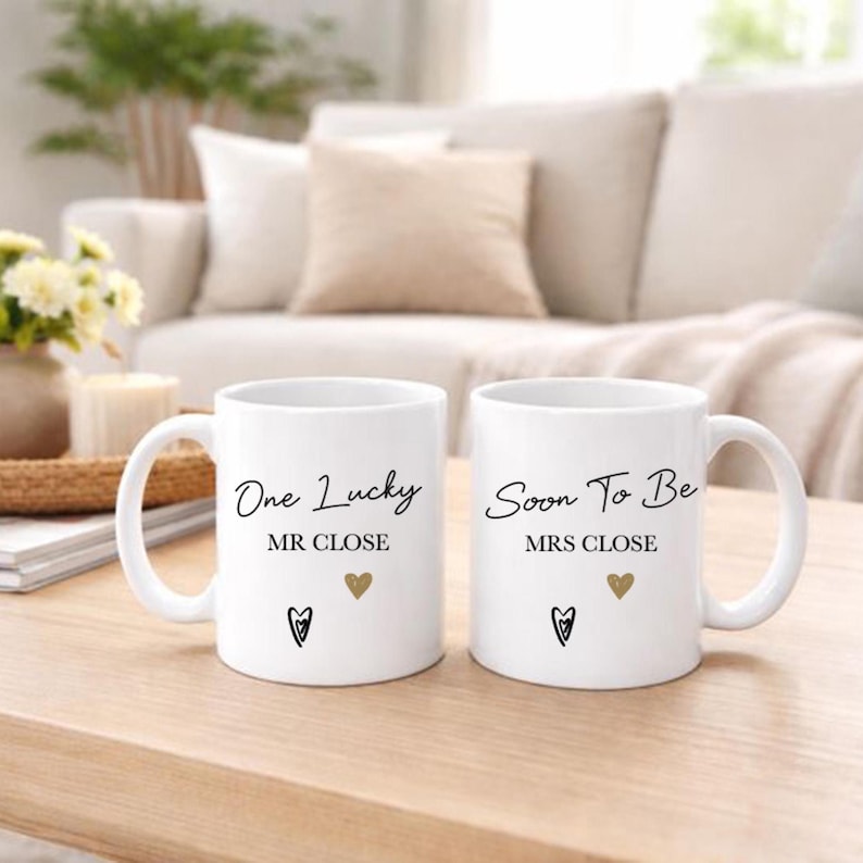 May include: Two white ceramic mugs with black script that reads "One Lucky MR CLOSE" and "Soon To Be MRS CLOSE". Each mug has a gold heart and a black heart. The mugs are on a wooden table.