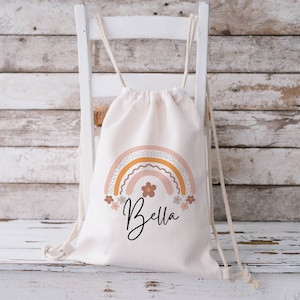 May include: A white canvas drawstring bag with a rainbow design in shades of pink, orange, and yellow. The rainbow has a dotted pattern and is surrounded by small flowers. The bag has the name "Bella" printed in black below the rainbow.