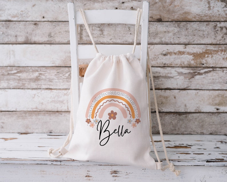 May include: A white canvas drawstring bag with a rainbow design in shades of pink, orange, and yellow. The rainbow has a dotted pattern and is surrounded by small flowers. The bag has the name "Bella" printed in black below the rainbow.
