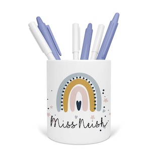 May include: White ceramic pencil holder with a rainbow design and the text "Miss Neish". The rainbow is in shades of yellow, orange, pink, and blue. There are small stars and hearts around the rainbow. The holder has five pens in it, four blue and one white.