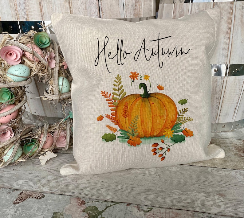 May include: A square beige throw pillow featuring the black cursive text Hello Autumn above a central illustration of an orange pumpkin surrounded by colorful fall foliage, including green leaves, orange wheat stalks, and small red berries.