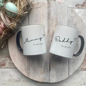 May include: Two white ceramic mugs with black handles and rims, inscribed with "Mummy" and "Daddy" in cursive, each with a heart symbol. The mugs are on a wooden surface, with an Easter wreath in the background.