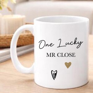 May include: White ceramic mug with the text "One Lucky MR CLOSE" in black script. The mug features a gold heart and a black heart design. The mug has a large handle and is suitable for hot or cold drinks.