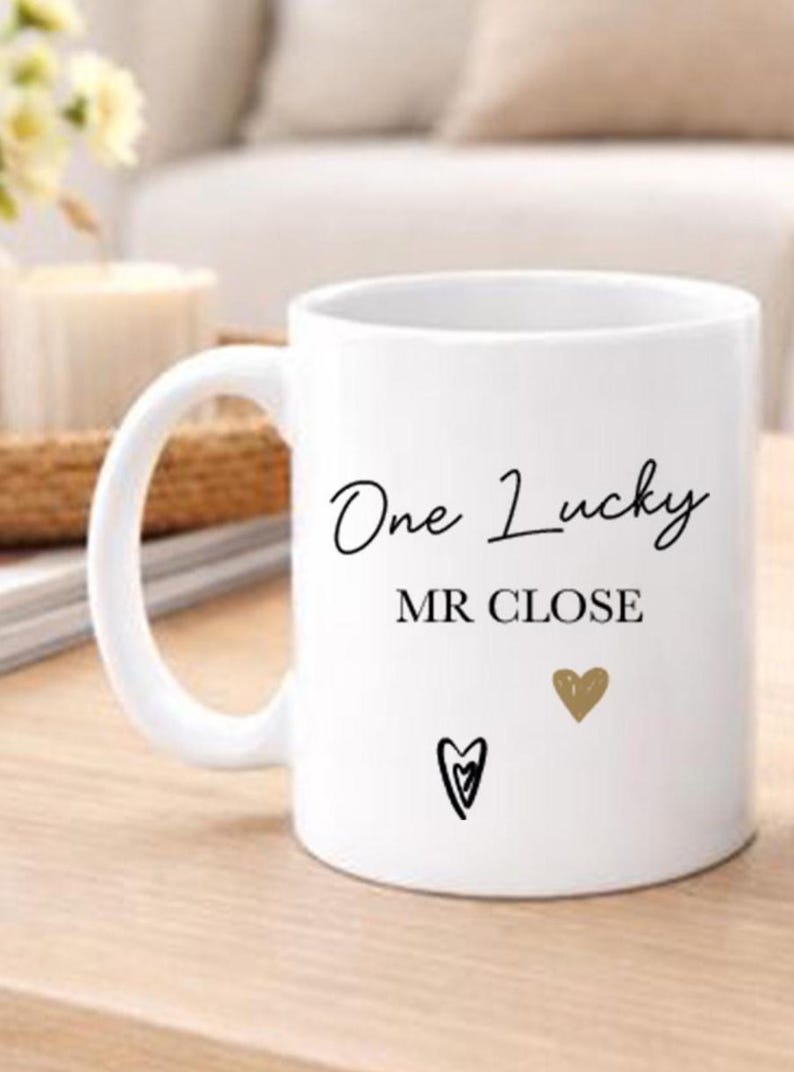 May include: White ceramic mug with the text "One Lucky MR CLOSE" in black script. The mug features a gold heart and a black heart design. The mug has a large handle and is suitable for hot or cold drinks.
