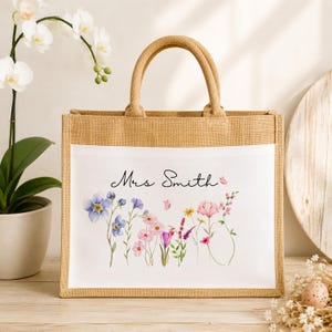 May include: A rectangular jute tote bag with two sturdy handles and a white front panel. The panel features a watercolor illustration of various wildflowers in shades of pink, purple, and blue, with the black cursive text Mrs Smith printed above.