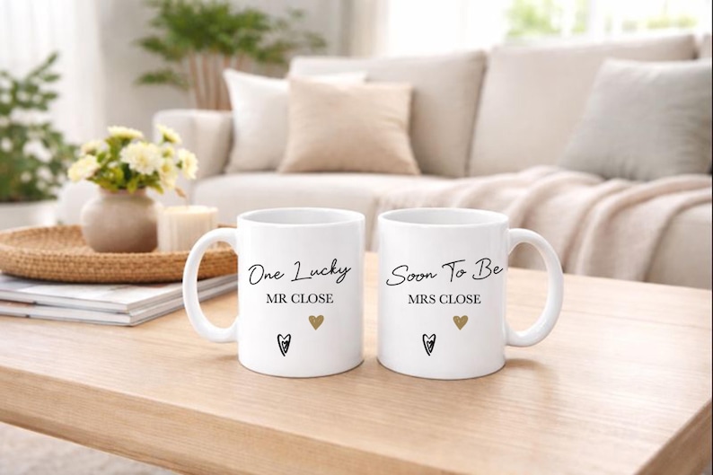 May include: Two white ceramic mugs with black script that reads "One Lucky MR CLOSE" and "Soon To Be MRS CLOSE". Each mug has a gold heart and a black heart-shaped design. The mugs are on a wooden table.