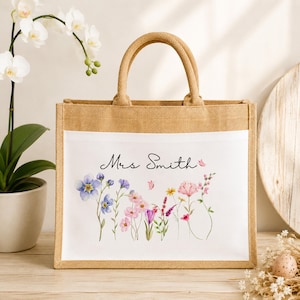 May include: A rectangular jute tote bag with two sturdy handles and a white front panel. The panel features a watercolor illustration of various wildflowers in shades of pink, purple, and blue, with the black cursive text Mrs Smith printed above.