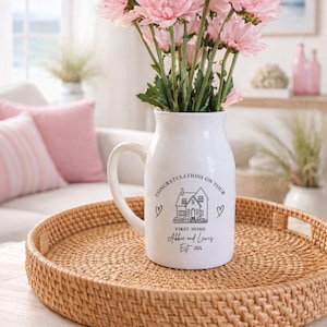 May include: A white ceramic pitcher filled with pink flowers, featuring a black printed graphic of a house, two hearts, and the text: CONGRATULATIONS ON YOUR FIRST HOME, Abbie and Lewis, Est 2026. The pitcher sits on a round woven rattan tray.