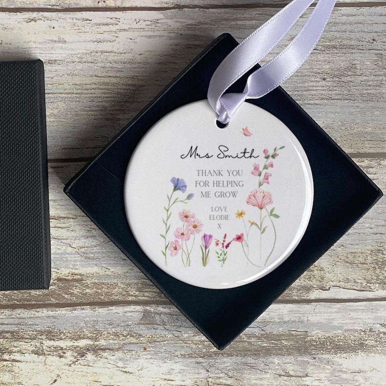May include: A round white ceramic ornament featuring a light purple ribbon for hanging. The surface displays colourful watercolour wildflowers and the text: Mrs Smith, THANK YOU FOR HELPING ME GROW, LOVE ELODIE x. The ornament is inside a black box.