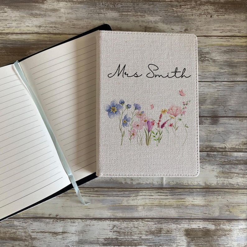 May include: A notebook with the name "Mrs Smith" in script on the cover. The cover has a floral design with blue, pink, and purple flowers. The notebook is open, showing lined pages and a blue ribbon.