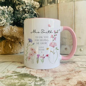 May include: A white ceramic mug with a pink handle and rim, featuring a colourful floral print and black text that reads: Mrs Smith, THANK YOU FOR HELPING ME GROW, LOVE ELODIE x. The design includes various delicate wildflowers and a small butterfly.