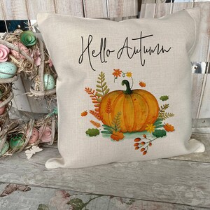 May include: A square beige throw pillow featuring the black cursive text Hello Autumn above a central illustration of an orange pumpkin surrounded by colorful fall foliage, including green leaves, orange wheat stalks, and small red berries.