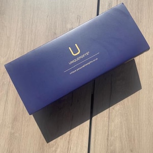 May include: A navy blue rectangular gift box with the text "UNIQUEGIFTS" and the letter "U" in gold. The website address "unique-personalisedgifts.co.uk" is printed below. The box is on a light brown wooden surface.