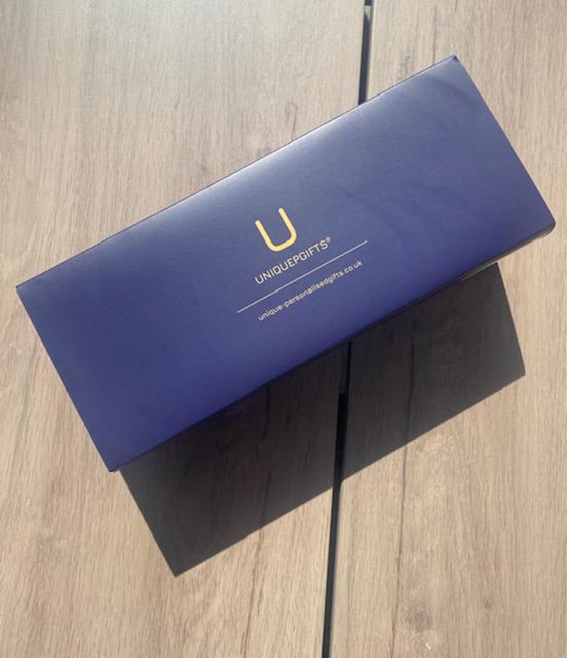 May include: A navy blue rectangular gift box with the text "UNIQUEGIFTS" and the letter "U" in gold. The website address "unique-personalisedgifts.co.uk" is printed below. The box is on a light brown wooden surface.