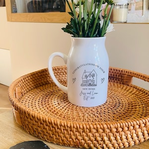 May include: A white ceramic pitcher filled with white daisies, featuring black text that reads CONGRATULATIONS ON YOUR NEW HOME, Jess and Liam, Est 2025, and a line drawing of a house. The pitcher sits on a round woven rattan tray.