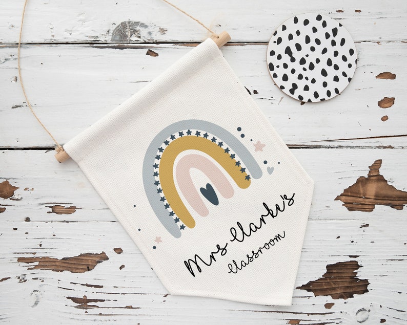 May include: A cream-colored classroom banner with a rainbow design in muted colors, including gold, blue, and pink. The banner has the text "Mrs. Clarke's classroom" in a handwritten font. A round coaster with black spots is also visible.