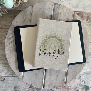 May include: A beige passport holder featuring a sage green rainbow graphic surrounded by white flowers and the text Miss Ward in black script. The holder is positioned on top of a black notebook with cream-coloured pages, displaying a textured fabric finish.