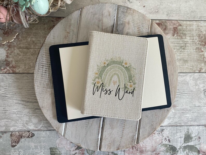 May include: A beige passport holder featuring a sage green rainbow graphic surrounded by white flowers and the text Miss Ward in black script. The holder is positioned on top of a black notebook with cream-coloured pages, displaying a textured fabric finish.