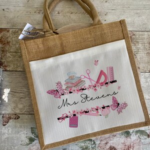 May include: A brown burlap tote bag with a white fabric insert. The insert features a pink and black design with butterflies, books, scissors, a ruler, a pencil, a calculator, and the text "Mrs. Stevens".