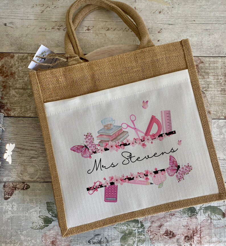 May include: A brown burlap tote bag with a white fabric insert. The insert features a pink and black design with butterflies, books, scissors, a ruler, a pencil, a calculator, and the text "Mrs. Stevens".