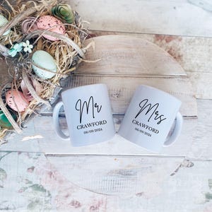 May include: Two white ceramic mugs with black lettering. The mug on the left says "Mr. Crawford 06.09.2024" and the mug on the right says "Mrs. Crawford 06.09.2024".