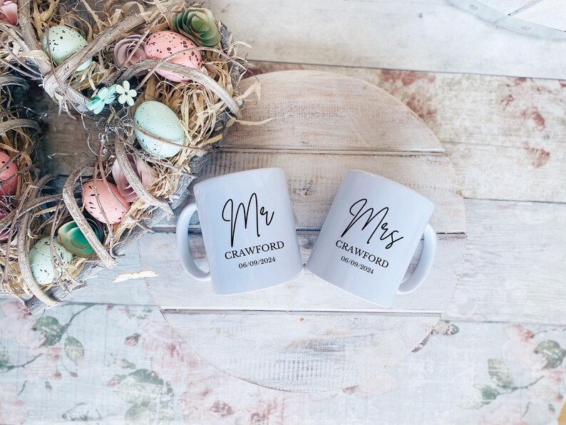 May include: Two white ceramic mugs with black lettering. The mug on the left says "Mr. Crawford 06.09.2024" and the mug on the right says "Mrs. Crawford 06.09.2024".