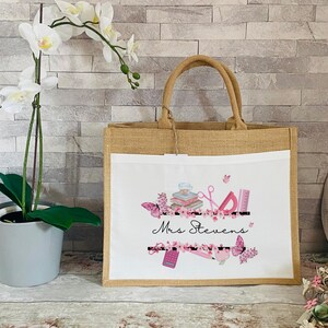 May include: A white canvas tote bag with brown jute handles. The bag has a pink and white floral design with the text "Mrs. Stevens" and school supplies.