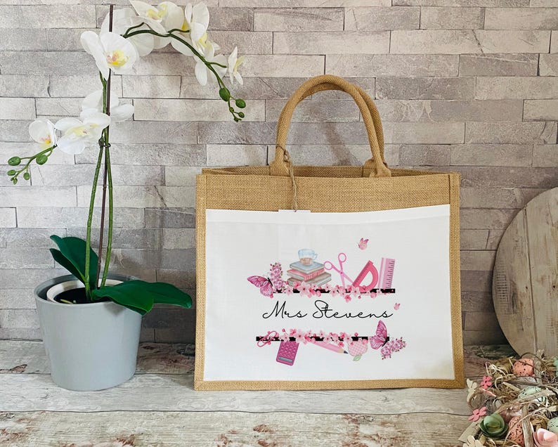May include: A white canvas tote bag with brown jute handles. The bag has a pink and white floral design with the text "Mrs. Stevens" and school supplies.