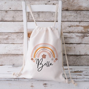 May include: A white canvas drawstring bag with a rainbow design in shades of pink, orange, and yellow. The rainbow has a dotted pattern and is surrounded by small flowers. The bag has the name "Bella" printed in black below the rainbow.