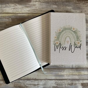 May include: A notebook with a textured off-white cover featuring a sage green rainbow graphic, white flowers, and the text Miss Ward in black script. The notebook is open to reveal lined white pages and a light blue ribbon bookmark attached to the spine.