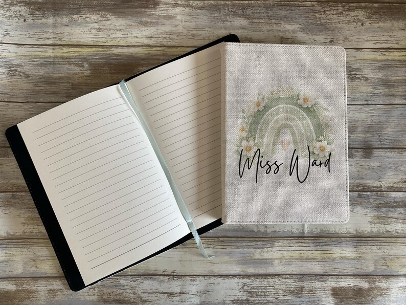 May include: A notebook with a textured off-white cover featuring a sage green rainbow graphic, white flowers, and the text Miss Ward in black script. The notebook is open to reveal lined white pages and a light blue ribbon bookmark attached to the spine.