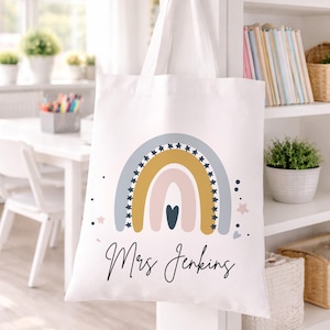 May include: White tote bag with a rainbow design in pastel colors, including blue, yellow, and pink, with star accents. The name "Mrs Jenkins" is written in cursive script below the rainbow. The bag is hanging on a white wall.
