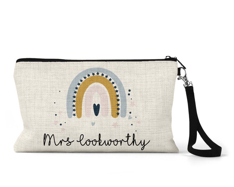 May include: A white canvas makeup bag with a black zipper and a black strap. The bag features a rainbow design with stars and a heart in the centre. The text "Mrs. Lookworthy" is printed on the bag.