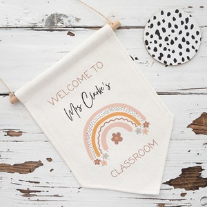 K&ouml;nnte beinhalten: Ein wei&szlig;es Canvas-Banner mit dem Text "Willkommen in der Klasse von Frau Clarke" und einem Regenbogen mit Blumen. Das Banner h&auml;ngt an einer Holzleiste.