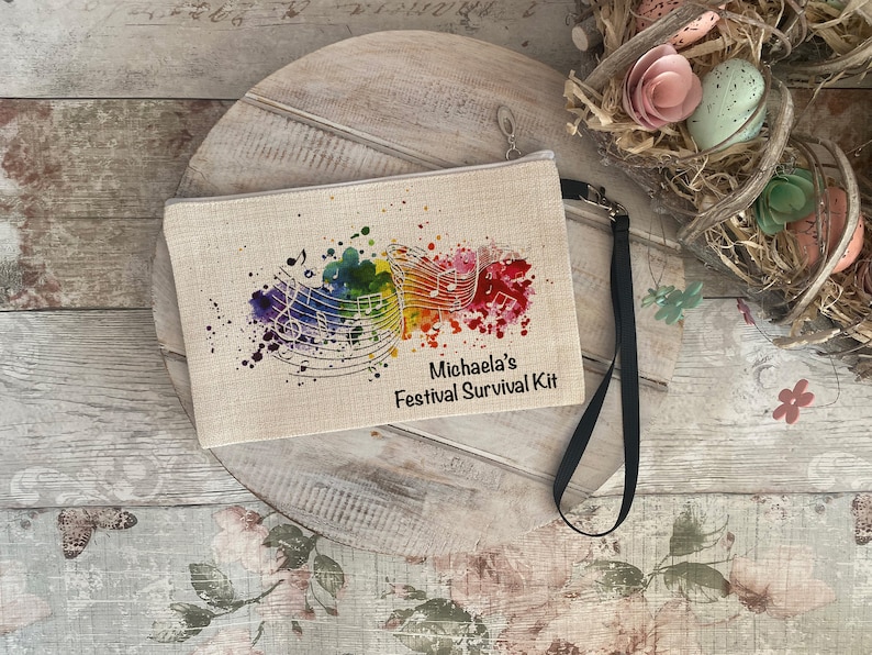 Op de afbeelding: Een beige canvas tasje met een zwarte rits en een zwarte band. Het tasje heeft een kleurrijk aquarel ontwerp met muzieknoten en de tekst "Michaela's Festival Survival Kit".
