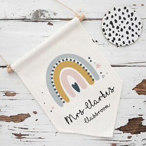 May include: A cream-colored classroom banner with a rainbow design in muted colors, including gold, blue, and pink. The banner has the text "Mrs. Clark's classroom" in a handwritten font. A round coaster with black spots is also visible.