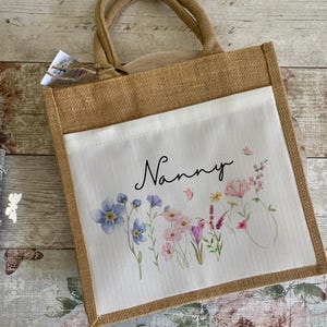 K&ouml;nnte beinhalten: Eine braune Jute-Tragetasche mit einem wei&szlig;en Stoffeinsatz. Der Einsatz zeigt ein Blumenmuster mit rosa, lila und blauen Blumen und dem Wort "Nanny" in schwarzer Schrift.